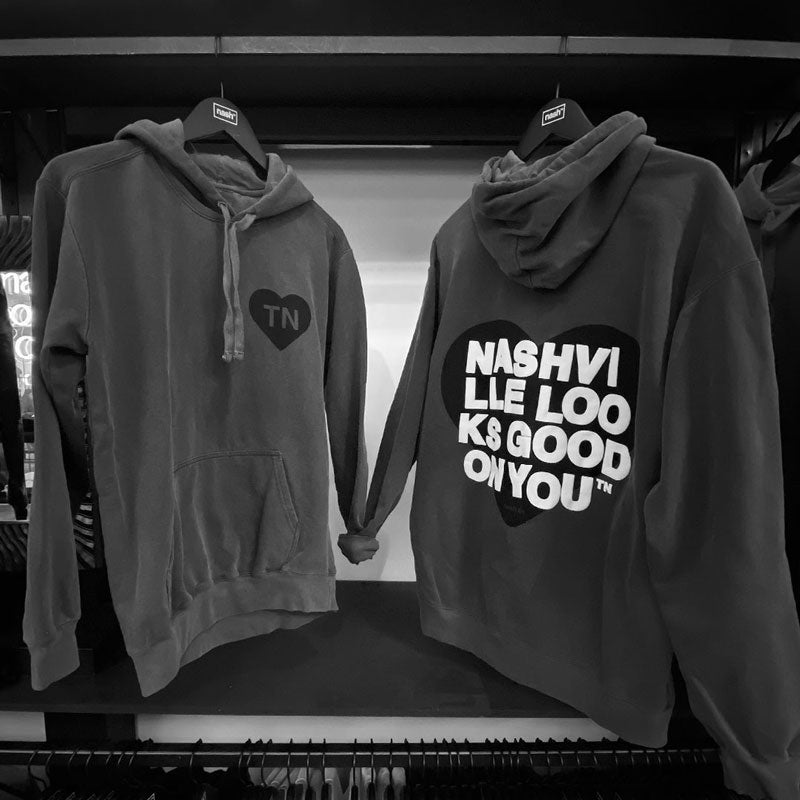 Back and front sides of gray hooded sweatshirt hanging on a rack. The sweatshirts are tied into a knot at the wrist cuff to make it look like they are holding hands. Backside has a black heart with white text printed on top that reads nashville looks good on youᵀᴺ in wrapped text. The front side of hoodie has a small black heart with letters TN on chest. TN is the abbreviation of Tennessee.
