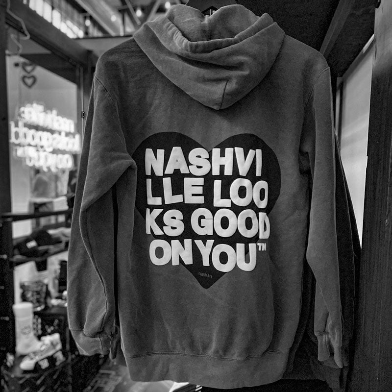 Back side of gray hooded sweatshirt with retail store in background. Backside has a black heart with white text printed on top that reads nashville looks good on youᵀᴺ in wrapped text. 