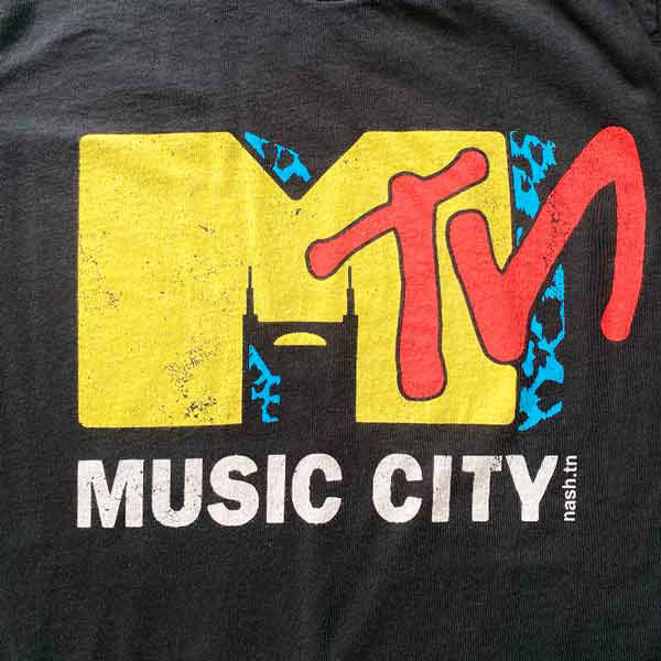 Closeup look at the colorful design on black T-shirt. Nashville is known as Music City and this shirt is a parody of a vintage design. The Batman Building is shown in the negative space of the yellow letter M. TN is written in red at an angle across the M. Music City is written in white block text across the bottom of the design.