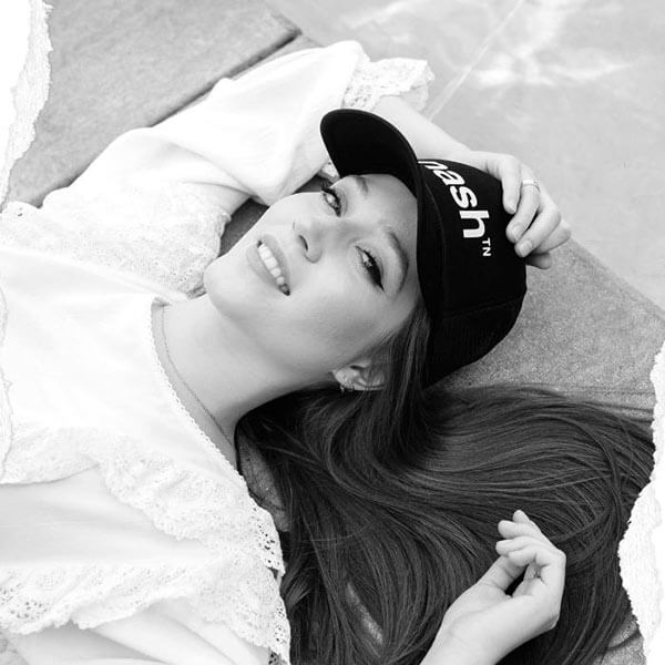Woman in white shirt lying on the ground. Her arms are up near her head. She's slightly smiling looking at the camera and wearing a black baseball cap. earing Nashville Trucker Hat