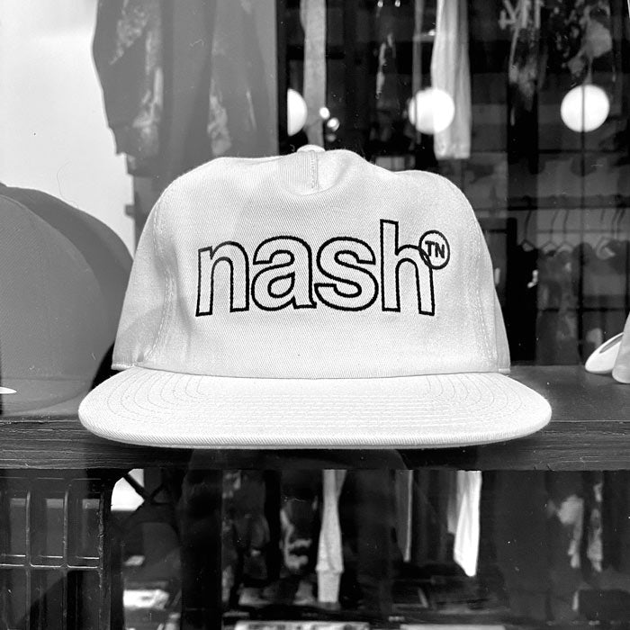 White hat with black outlined letters on display shelf with a blurry retail store background. Nashᵀᴺ is outlined in black and the superscript TN is inside a black, outlined circle.