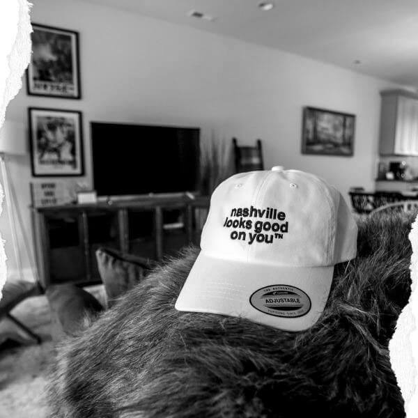 White baseball cap with black letters on a furry surface with living room items in the background.  The white baseball cap has black letters and says nashville looks good on youᵀᴺ on the front. There is a silver and black oval sticker on the visor.   