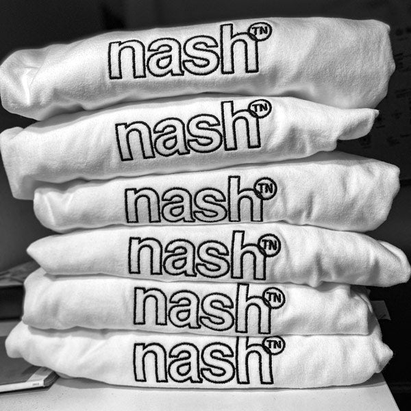 There is a folded stack of 6 white shirts on a flat surface. The black outline logo, nashᵀᴺ,  is facing out.