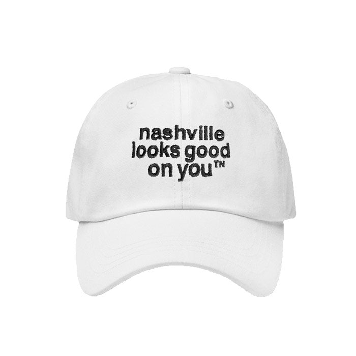 White baseball cap with black letters on a white background. Hat is angled slightly to the left and has nashville looks good on youᵀᴺ embroidered in black on the front of the cap. The visor is slightly curved and there is a silver and black oval shaped sticker on the visor that says Authentic Classics.
