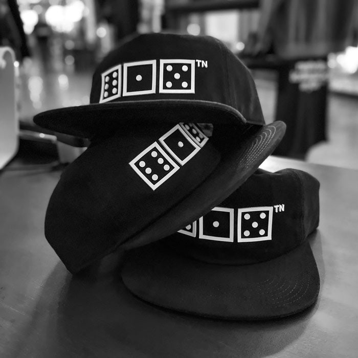There are three black baseball caps stacked on each other in a wobbly stack. The baseball hats are the same design. Black hats with white squares and dots. There is a small TN at the top right corner of the design. The TN looks like a trademark symbol, but this is a design mark for Tennessee. The squares look like game dice with the numbers six, one, five. 615 is the area code for Nashville, Tennessee, USA.