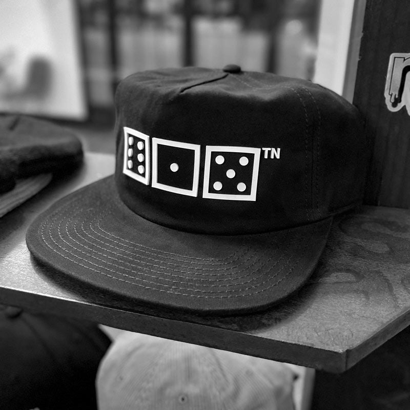 Black baseball hat with flat bill shown on a store shelf. The hat has a white design of squares and dots and a small TN at the top right corner. The squares are meant to look like game dice showing the numbers six, one, five. 615 is the area code for Nashville, Tennessee. 