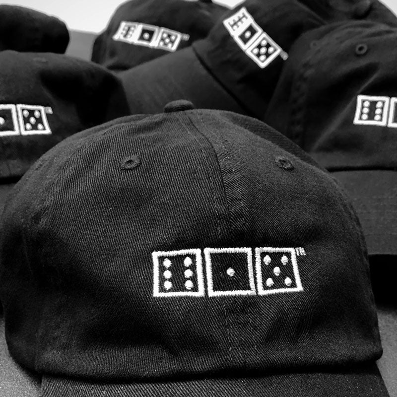 Closeup of black baseball cap and several other hats in the background. White embroidered game dice on the front of the hats with six, one, and five. 615 is the area code for Nashville, Tennessee.