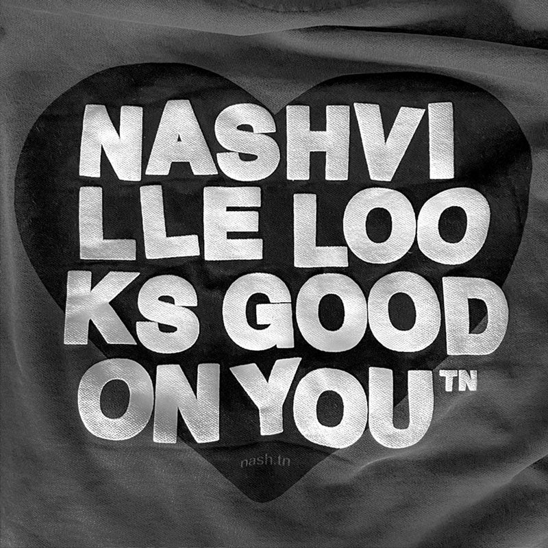 Close up detail of the back side design of the gray hooded sweatshirt. There is a black heart with white words across the heart. The white letters have a raised, puff, appearance and spell out nashville looks good on youᵀᴺ in wrapped text. 