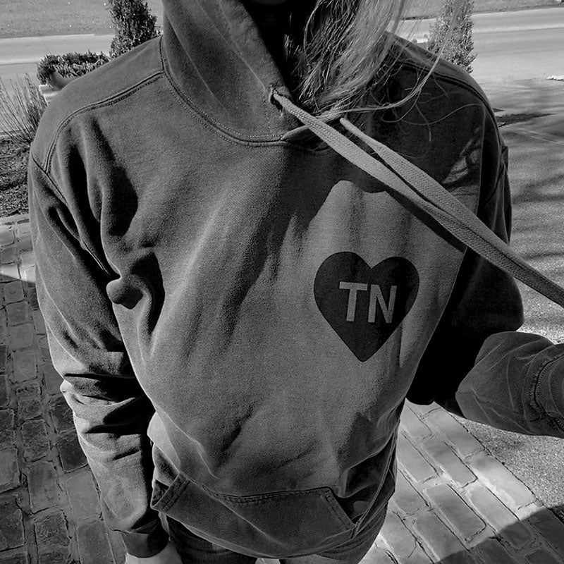Person wearing a gray hooded sweatshirt pulling the hood strings out to the side. There is a black heart with the letters TN on the front, left chest side. TN is the abbreviation for Tennessee.