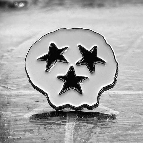 A close up of the white enamel, skull shaped fashion pin. This pin has three black stars that represent both Tennessee's Tri-Star moniker and a skeleton's face.
