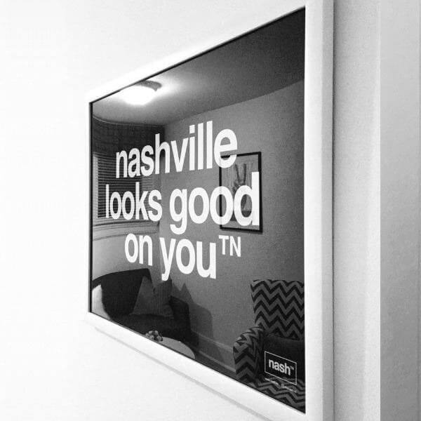 Angled view of black rectangle poster print framed in white. There is a reflection of furniture in the glass. The framed print is the nashᵀᴺ  slogan, nashville looks good on youᵀᴺ printed in white.