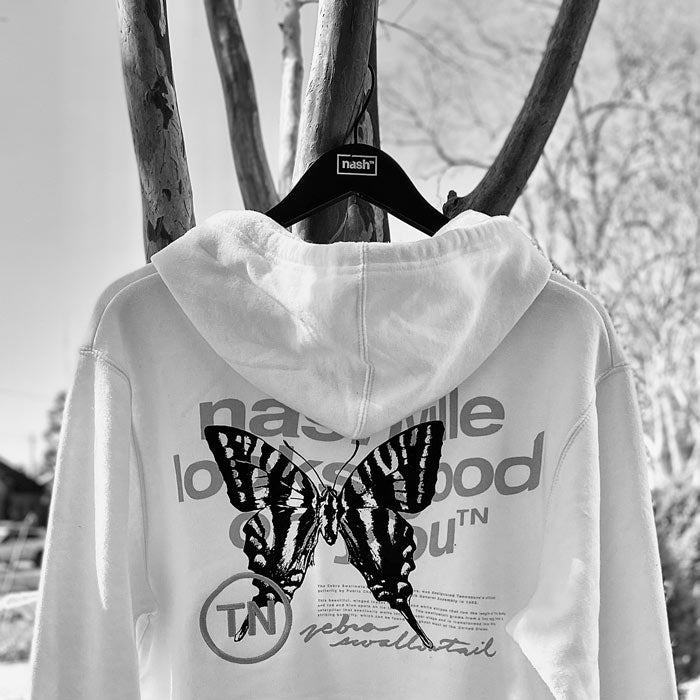 Black and white image showing backside of sweatshirt hoodie hanging from a tree on a black coat hanger. There is a large swallowtail butterfly image in black. There are words surrounding the butterfly that read: nashville looks good on youᵀᴺ  in gray wavy text, small block text about the butterfly, zebra swallowtail in cursive writing and the letters TN inside a circle.