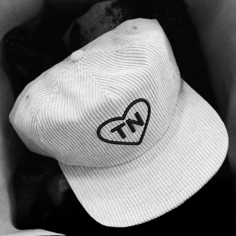 Stylized image of products in a shopping bag. The item on top is a gray corduroy baseball cap. The hat has a black heart and TN on the front. 