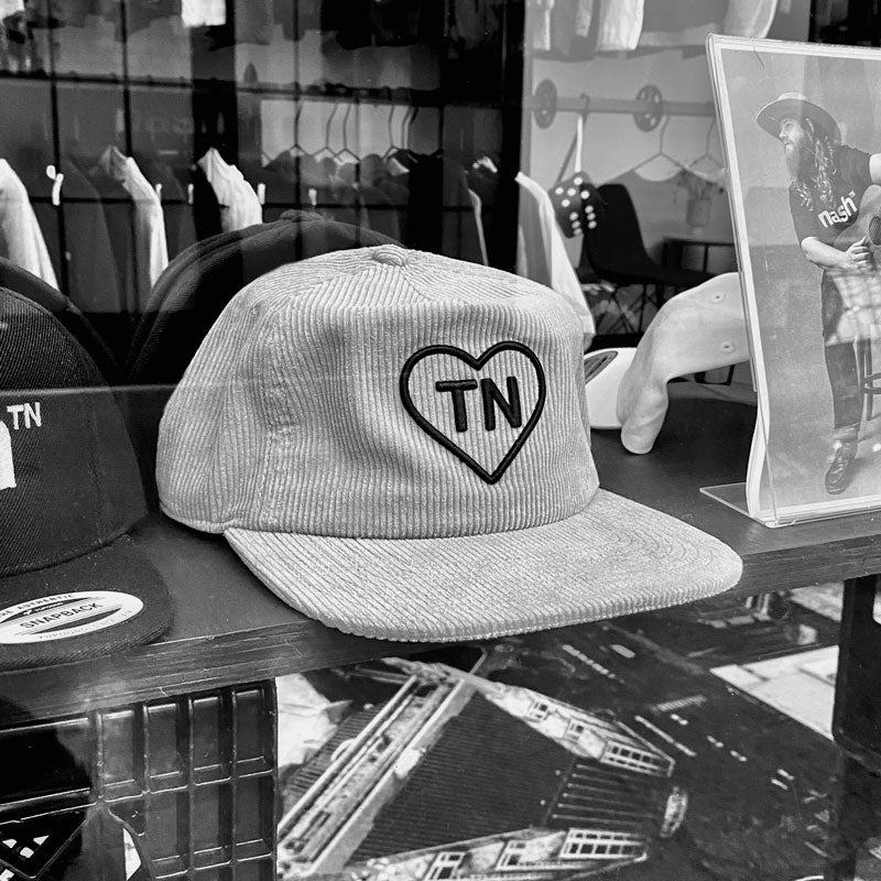 Gray corduroy baseball cap on a display shelf. The hat has a black outlined heart design on the front with the letters TN inside the heart. TN is the abbreviation for Tennessee. 