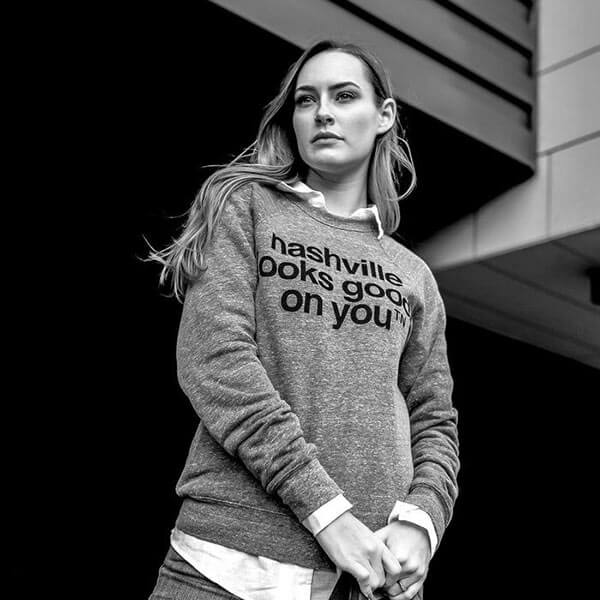 Woman wearing gray crew neck sweatshirt with black lettering and white button down shirt underneath in front of a dark colored house. Her hands are clasped in front of her at the hip.