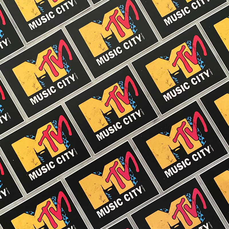 Rows of Mᵀᴺ stickers. Nashville is known as Music City.  Orange M for Music City and red TN for Tennessee.  Music City in white block text under the letter M. The Batman Building is shown in the negative space of the M. 