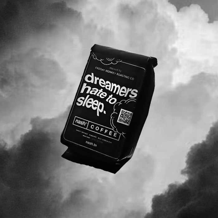Black coffee bag shown on a cloud background. The bag label has white text and cloud shape outlines. The text is wavy and says dreamers hate to sleep. 