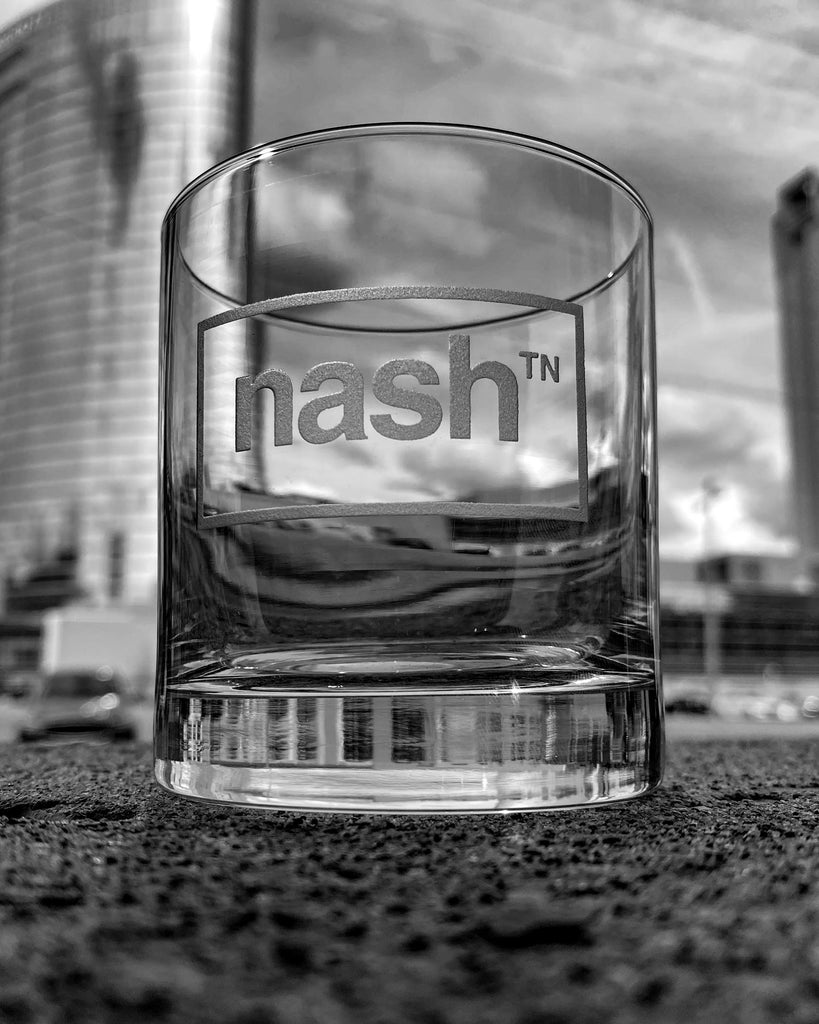 Clear rocks glass with frosted letters on a gray blurry background. On the front center of the glass, nashᵀᴺ is etched inside of a horizontal rectangle.