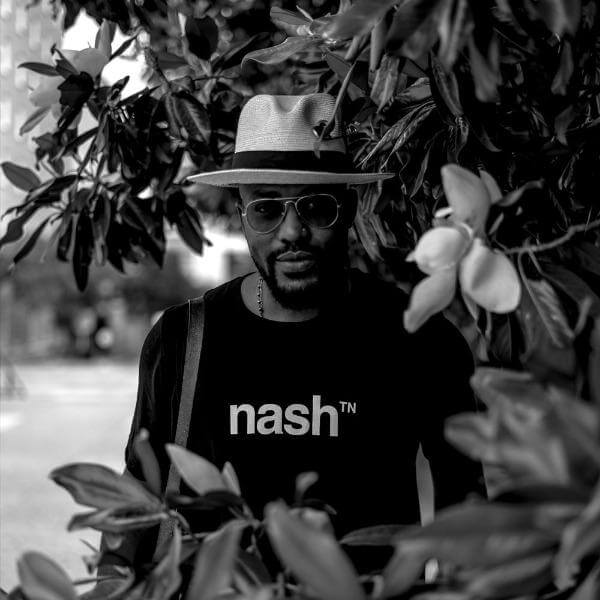 Man with hat and sunglasses standing among Magnolia tree branches is wearing a black, short sleeved crewneck T-shirt with white nashᵀᴺ  logo on the front.
