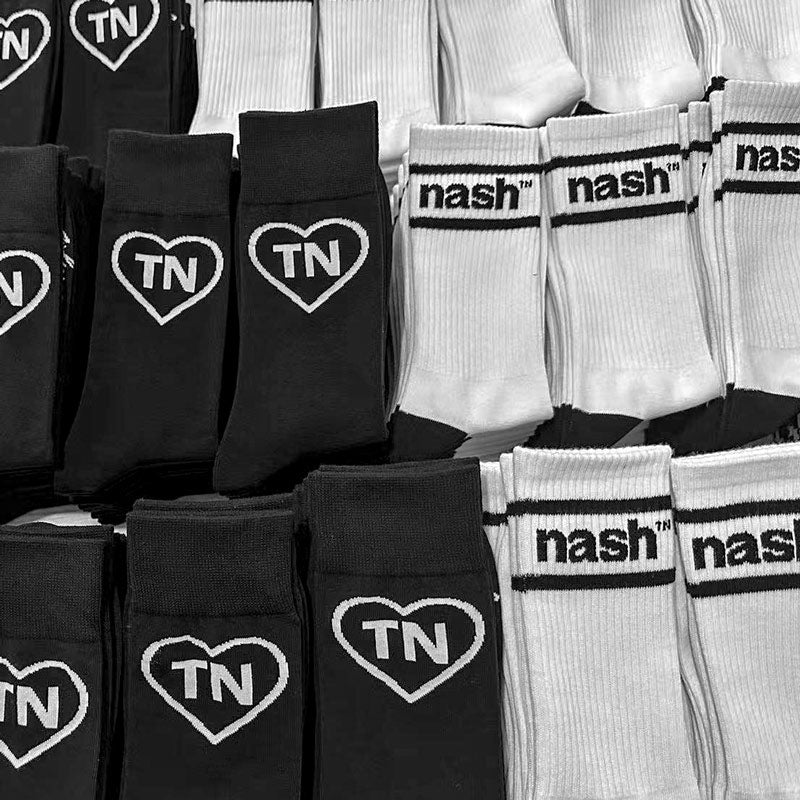 Stacks of folded black and white pairs of socks. The black socks on the left show a white heart outline and the letters TN inside the heart. The socks on the left show nashᵀᴺ  in black with a black stripe above and below the logo.