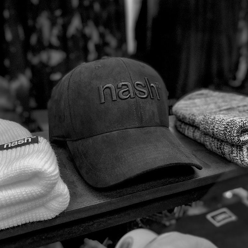 Black faux suede baseball cap shown on shelf with other products. The hat has black embroidery on the front that reads nash with a small TN at the top right. TN is the abbreviation for Tennessee and looks like the trademark symbol.