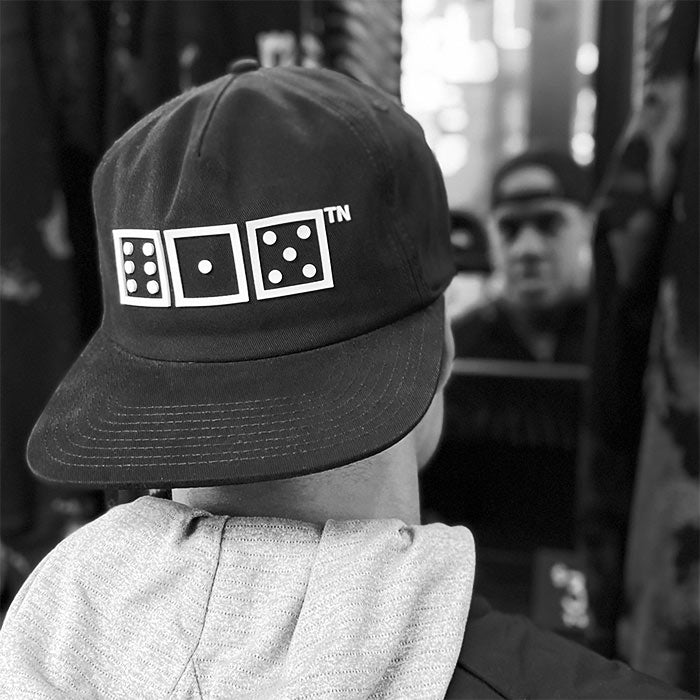 A man  who is wearing a baseball cap backward is looking at himself in mirror. He is wearing a black baseball hat that has squares and dots. There is a small TN at the top right corner of the design. The squares look like game dice with the numbers six, one, five. 615 is the area code for Nashville, Tennessee. 