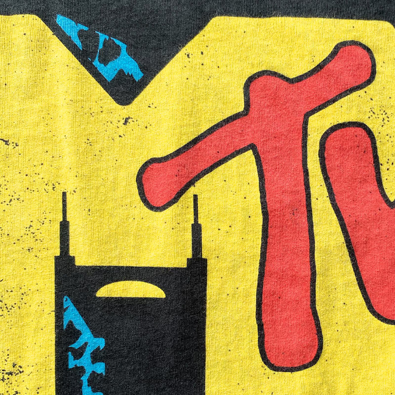 A closer look at the black short sleeve T-shirt with colorful design. Nashville is known as Music City and this shirt is a parody of a vintage design. The Batman Building is shown in the negative space of the yellow letter M. The letter T is written in red at an angle across the yellow M. There are small accents of blue cheetah shown at this detail.