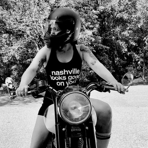 Woman on motorcycle wearing a helmet, black tank top, shorts and tall white socks with a stripe at the top. The tank top has white words on the front: nashville looks good on youᵀᴺ  