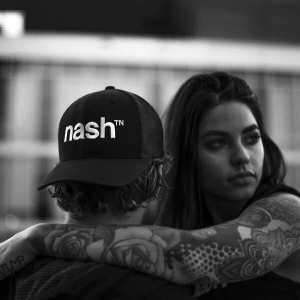 Man and woman on a blurry background. Man has his back to camera and wearing a black, backward baseball cap. The cap has the nashᵀᴺ  logo in white. The woman is facing the camera and has her arm around the man's neck draped across his shoulders. Her arm has a sleeve of tattoos in geometric shapes.
