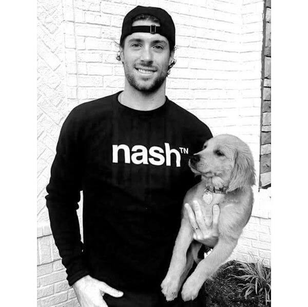 Man with beard and half smile is holding a puppy in front a white painted wall. He is wearing a backward, black hat and black crew neck sweatshirt with white nashᵀᴺ  logo.