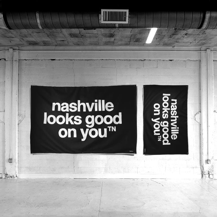 Two black canvases hang on a white painted block wall. The canvases have the phrase nashville looks good on youᵀᴺ painted in white. The large canvas is on the left and hangs horizontally. The small canvas hangs on the right and is hung vertically with the words reading sideways.    