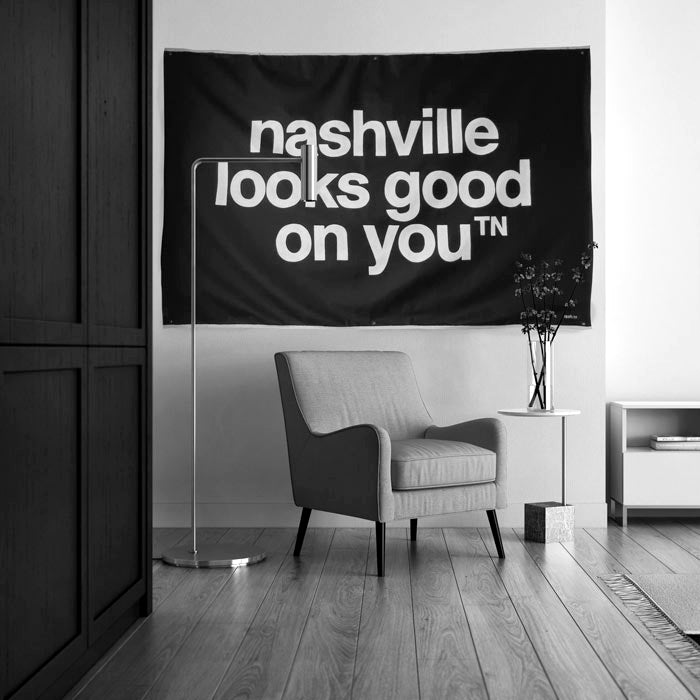 Black canvas with white letters hangs on a wall behind a chair and lamp. The canvas reads nashville looks good on youᵀᴺ  