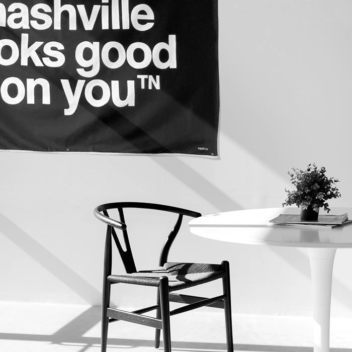Black canvas with white words hangs on a white wall above and to the left of a black chair and white table