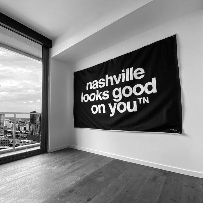 Black canvas with white letters hangs on a wall in an empty room. The canvas reads nashville looks good on youᵀᴺ. There is a large window on the left with city buildings in the distance.