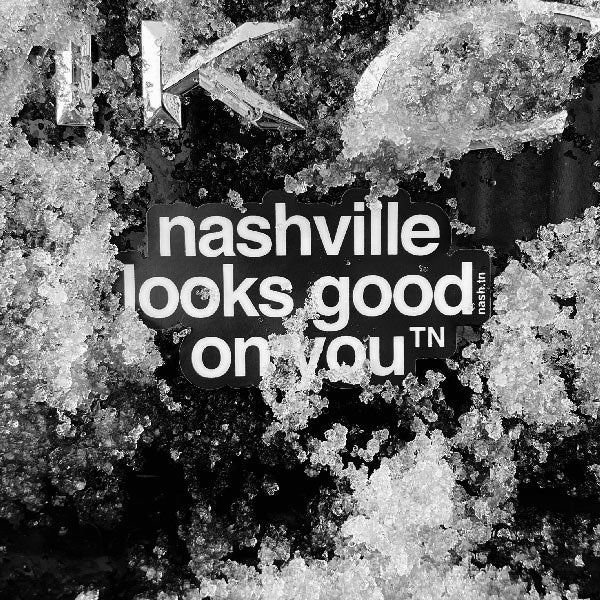 Sticker on the back of a car covered in snow. Black sticker with white letters on a dark background. The sticker says nashville looks good on youᵀᴺ with a small, superscript TN next to the letter u in you. TN is the abbreviation for Tennessee. Nashville is the capital city in Tennessee.