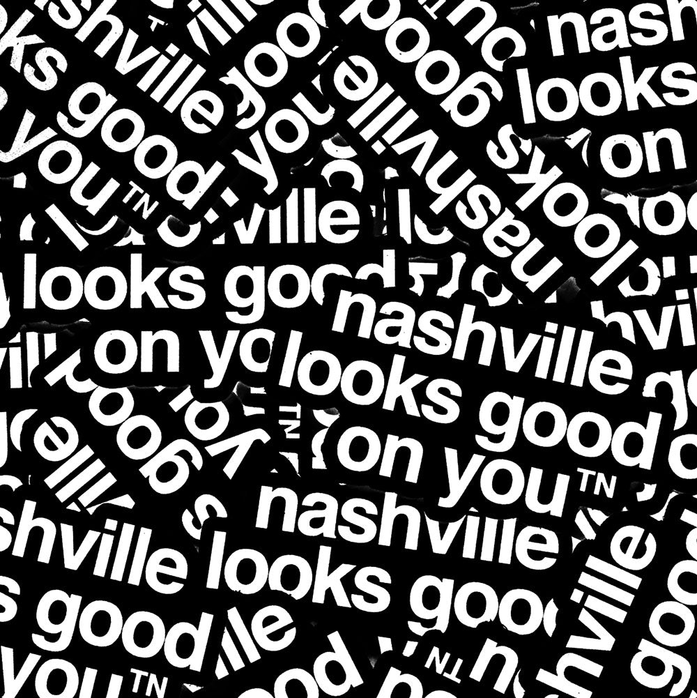 Pile of stickers on a table. Black stickers with white letters on a white background. The sticker says nashville looks good on youᵀᴺ with a small, superscript TN next to the letter u in you. TN is the abbreviation for Tennessee. Nashville is the capital city in Tennessee.