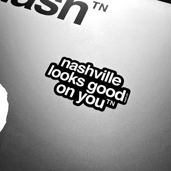 Black sticker with white letters on a metallic background. The sticker shape is cut around the letters. The sticker says nashville looks good on youᵀᴺ with a small, superscript TN next to the letter u in you. TN is the abbreviation for Tennessee. Nashville is the capital city in Tennessee.