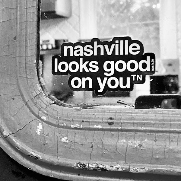 Black sticker with white letters on a mirror, the sticker shape is cut around the letters. The sticker says nashville looks good on youᵀᴺ with a small, superscript TN next to the letter u in you. TN is the abbreviation for Tennessee. Nashville is the capital city in Tennessee.