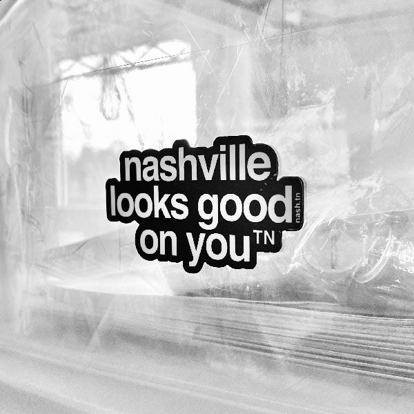 Black sticker with white letters on a hazy background, the sticker shape is cut around the letters. The sticker says nashville looks good on youᵀᴺ with a small, superscript TN next to the letter u in you. TN is the abbreviation for Tennessee. Nashville is the capital city in Tennessee.