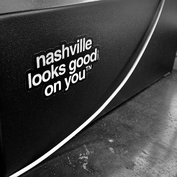 Black sticker with white letters stuck to the side of a black box. The sticker says nashville looks good on youᵀᴺ with a small, superscript TN next to the letter u in you. TN is the abbreviation for Tennessee. Nashville is the capital city in Tennessee.