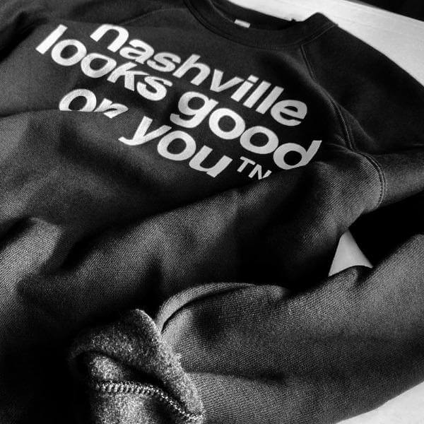 Angled view of black crew neck sweatshirt on a white surface. The sleeve is rolled up to show the fuzzy inside of the sweatshirt. The white text nashville looks good on youᵀᴺ is visible.  