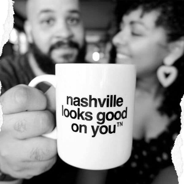 Blurry couple in the background, the lady has a heart shaped earring in her left ear. She is smiling and looking and the gentleman on her right side. He is holding a white coffee mug with black text close to the camera. Nashville looks good on youᵀᴺ is printed in black in the center of the mug.