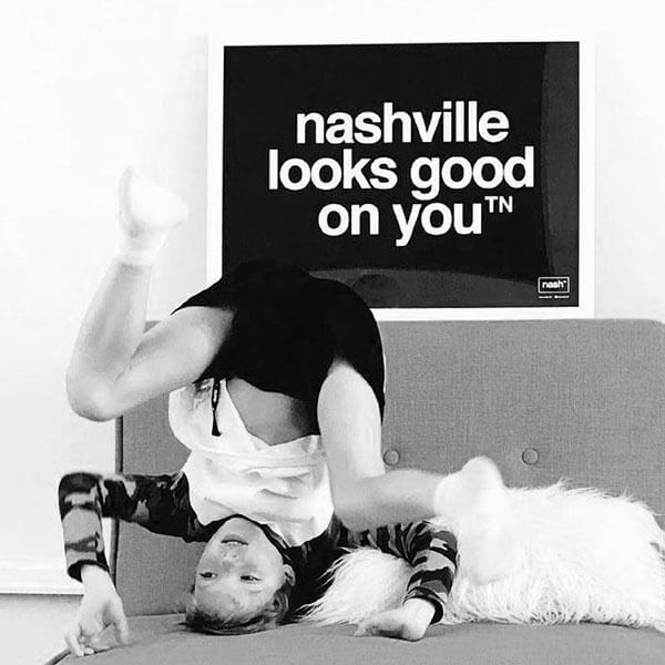 Black rectangle with white letters above a sofa with a kid playing upside down on the sofa below. The black rectangle is a poster and is framed in white. The poster has nashville looks good on youᵀᴺ printed in white.  