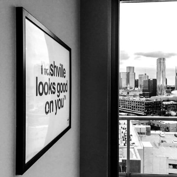 Angled view of white rectangle poster print framed in black. The framed print is the nashᵀᴺ slogan, nashville looks good on youᵀᴺ printed in black. On the right, in the distance there are large, skyscraper buildings.