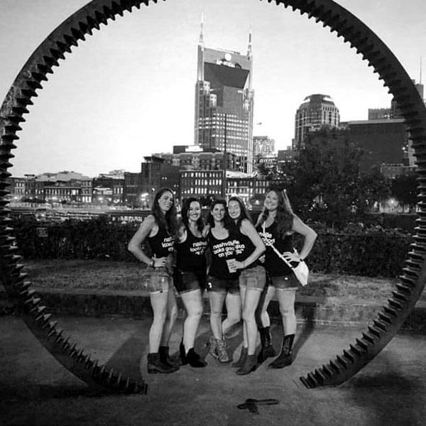 A group of ladies poses in front of the Nashville skyline wearing matching outfits: boots, shorts and black tank tops that read nashville looks good on you nashᵀᴺ  in white