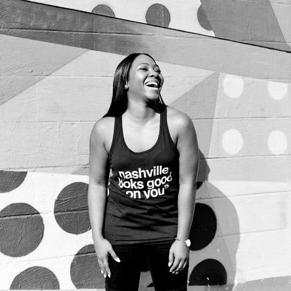 Woman laughing in front of a painted wall. She is wearing black pants and a black tank top with white words nashville looks good on youᵀᴺ  
