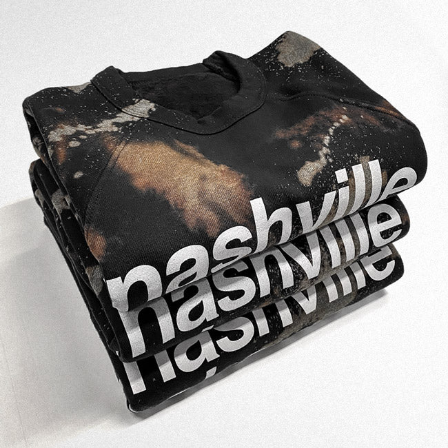 Three black reverse dyed sweatshirts in a stack on a white background. The sweatshirts have been dyed and bleached to have to have a marbled appearance with grays, browns and blues. White text is seen on each shirt and reads as nashville, nashville, nashville because of the way they are stacked.