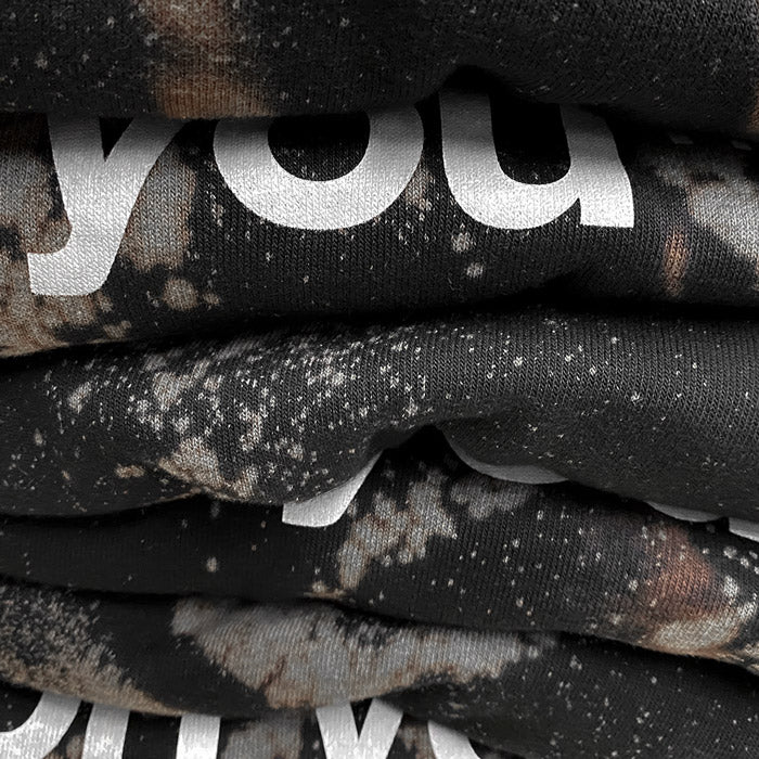Very closeup look at a stack of black reverse dyed sweatshirts. There are spots and streaks of gray and brown. There is obscured white text seen between the sweatshirt folds.