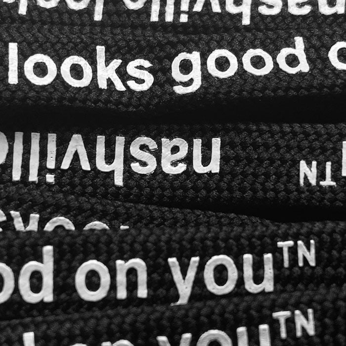 A close up look at the texture and printing on shoelaces. There are multiple black laces with white letters. On each lace, nashville looks good on youᵀᴺ is printed.
