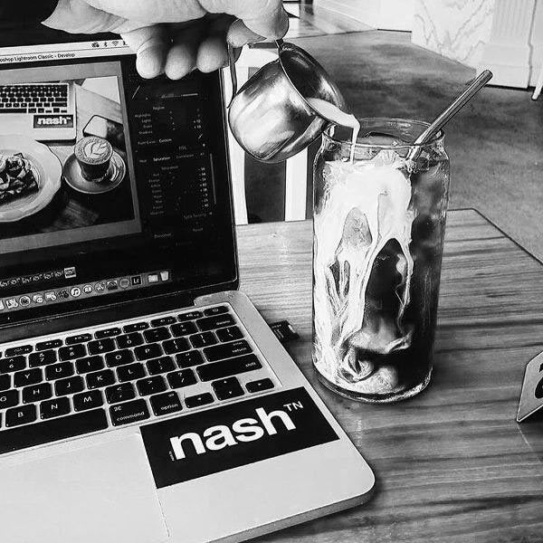 Black rectangle sticker with white nashᵀᴺ  logo affixed to laptop. Hand at the top of photo is pouring cream into a glass of coffee. 